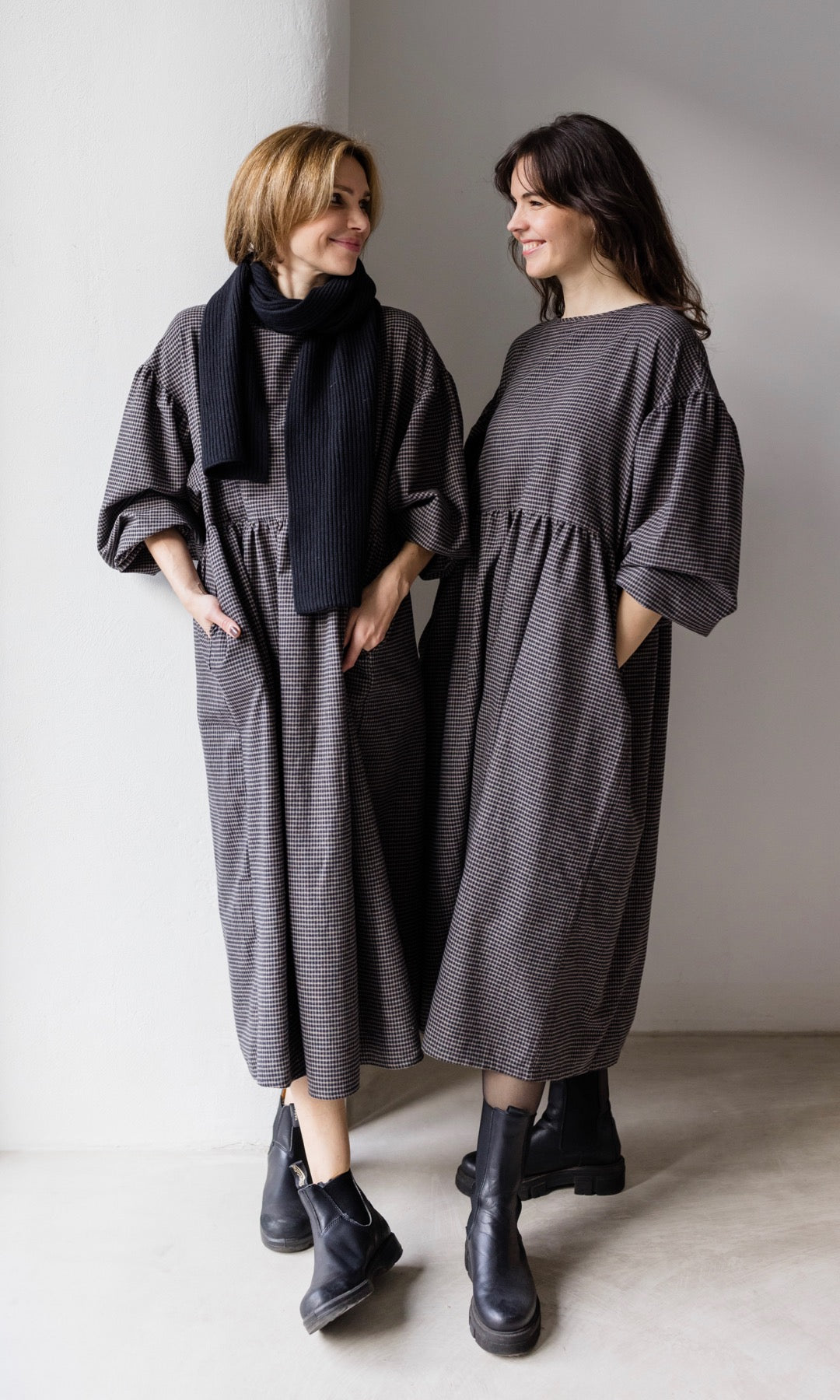 Winter Stillkleid Lovis in karo schwarz in zwei Looks mit schwarzen Stiefeln eine Variante mit schwarzem Schal Blicke seitlich zueinander Hände in Taschen