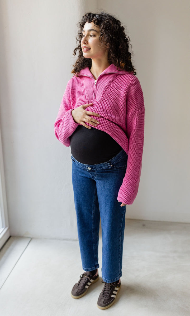 Schwangerschaftsjeans im French Fit Look in dunkelblau kombiniert mit rosa Strickpullover und braunen Sneakers. Inklusive elastischem Bauchbund. Blick seitlich lächelnd.
