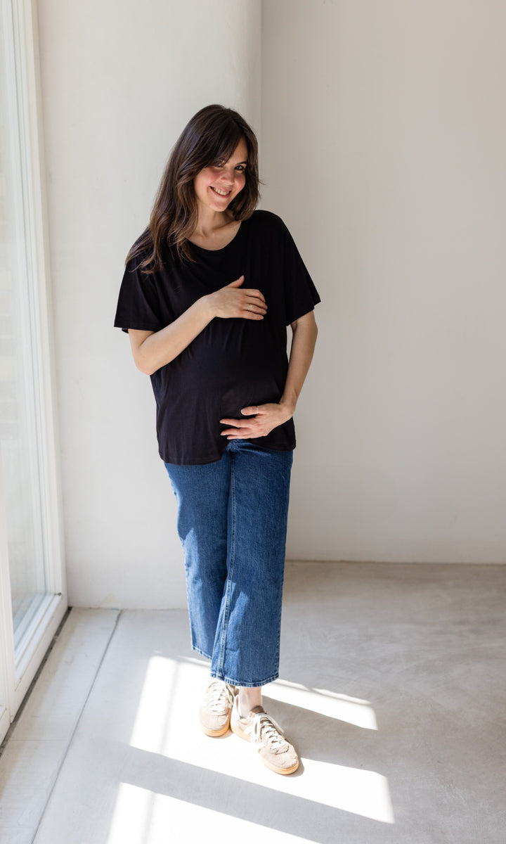 Schwangerschaftsjeans im French Fit Look in dunkelblau mit Bauchbund kombiniert mit schwarzem T-Shirt und braunen Sneakers. Blick gerade lachend.