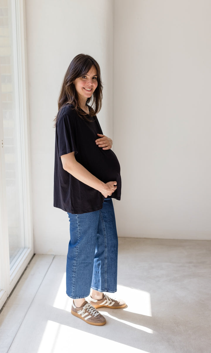 Schwangerschaftsjeans im French Fit Look in dunkelblau mit Bauchbund kombiniert mit schwarzem T-Shirt und braunen Sneakers. Blick seitlich lachend.
