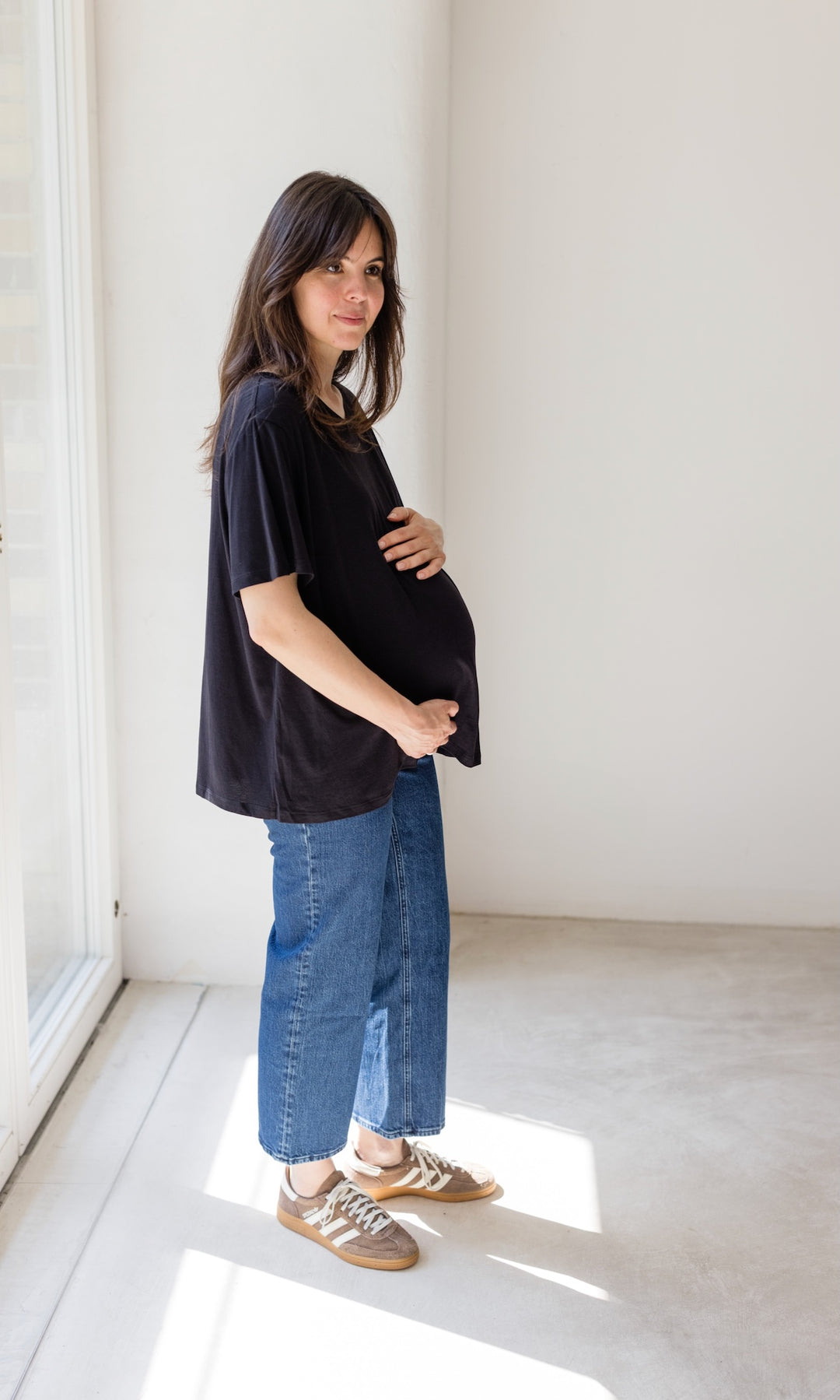 Schwangerschaftsjeans im French Fit Look in dunkelblau mit Bauchbund kombiniert mit schwarzem T-Shirt und braunen Sneakers. Blick seitlich.