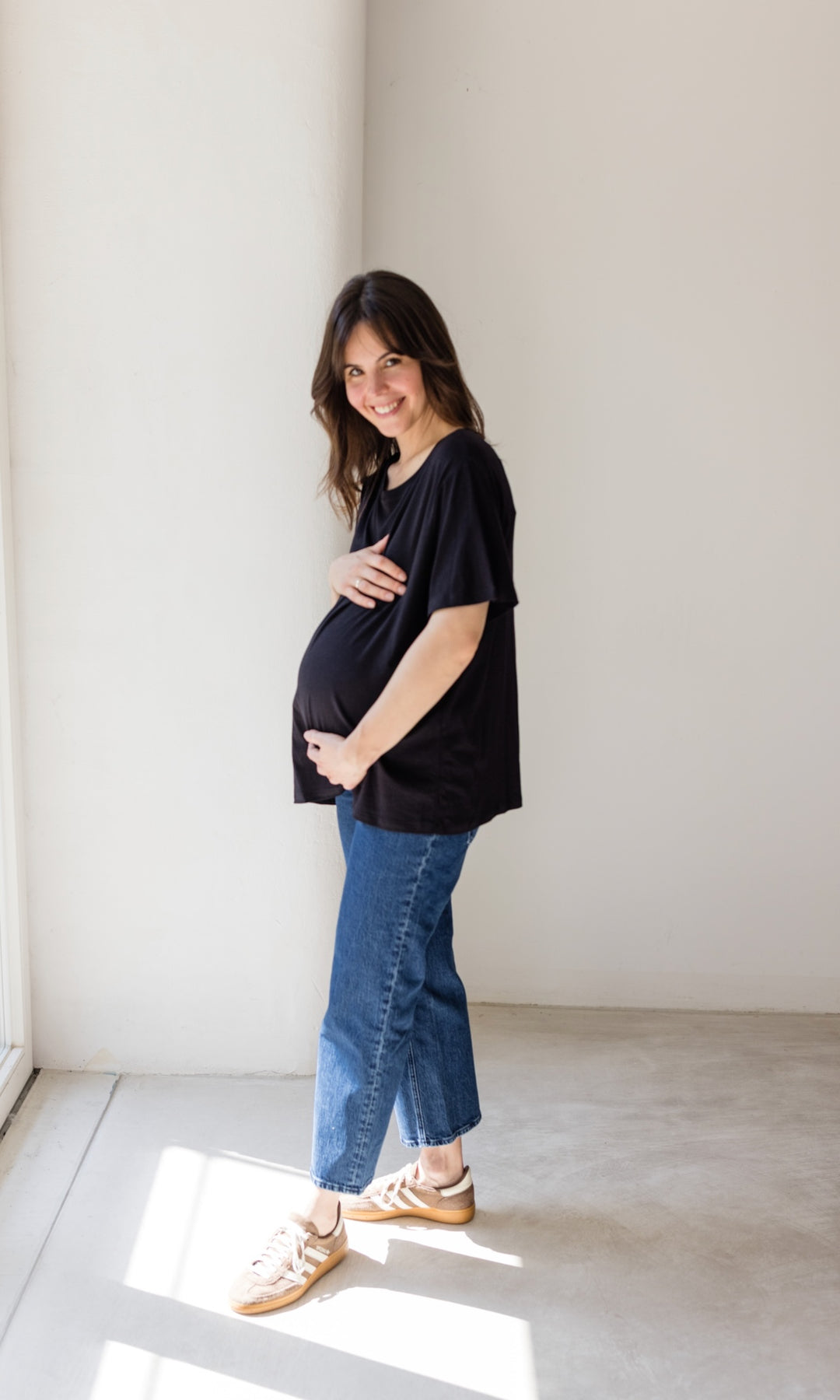 Schwangerschaftsjeans im French Fit Look in dunkelblau mit Bauchbund kombiniert mit schwarzem T-Shirt und braunen Sneakers. Blick seitlich lachend.