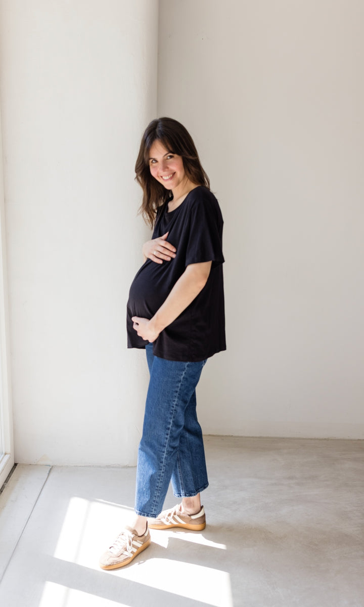 Schwangerschaftsjeans im French Fit Look in dunkelblau mit Bauchbund kombiniert mit schwarzem T-Shirt und braunen Sneakers. Blick seitlich lachend.