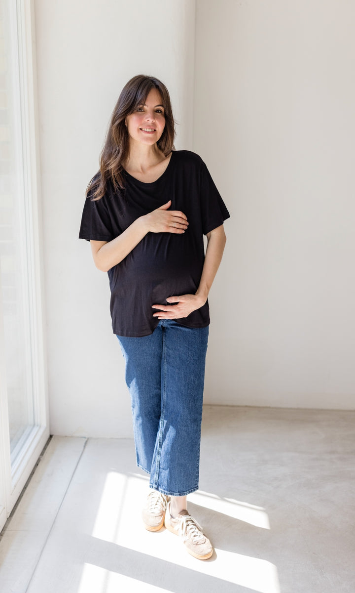 Schwangerschaftsjeans im French Fit Look in dunkelblau kombiniert mit schwarzem T-Shirt und braunen Sneakers. Blick gerade lachend.