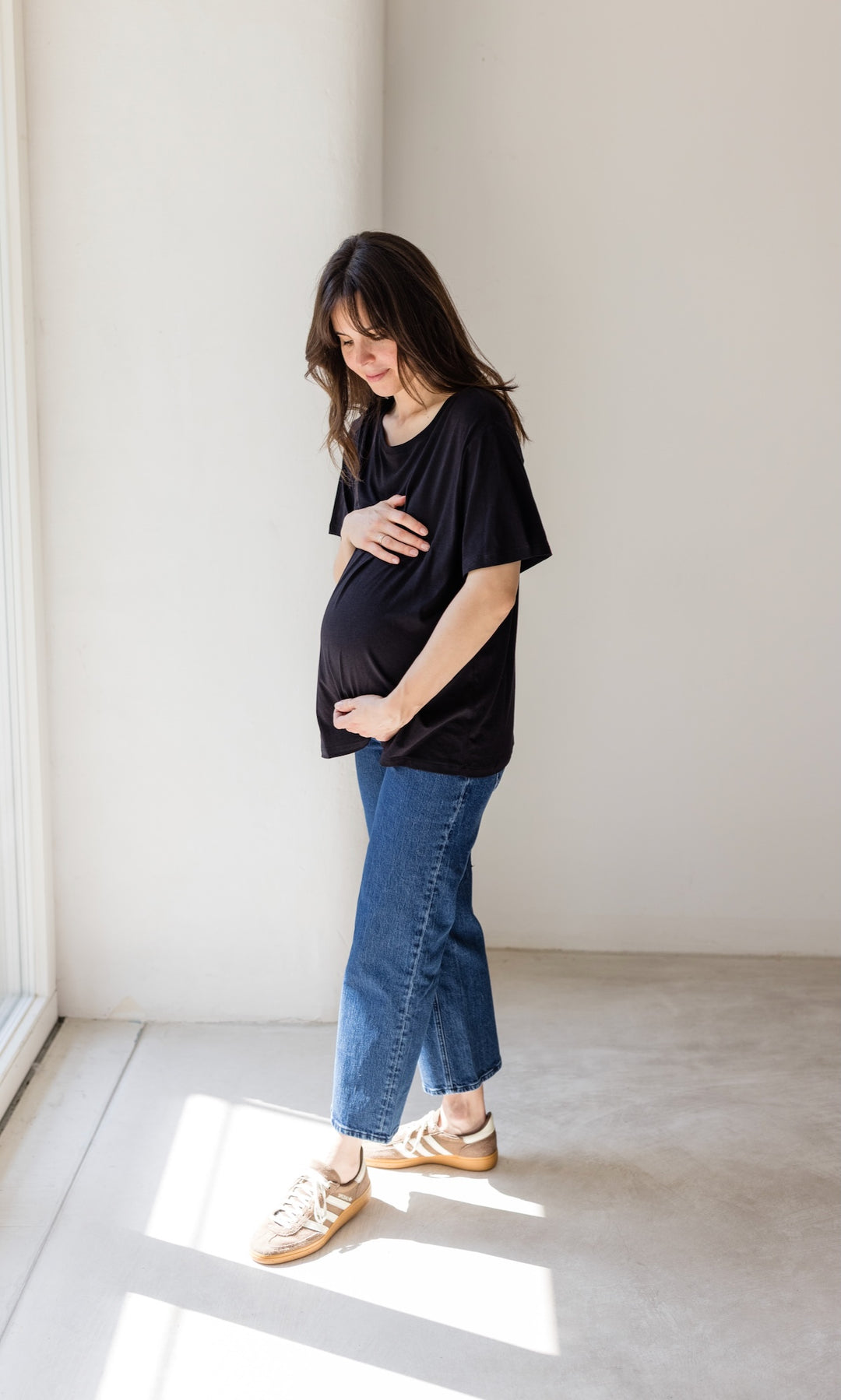 Schwangerschaftsjeans im French Fit Look in dunkelblau mit Bauchbund kombiniert mit schwarzem T-Shirt und braunen Sneakers. Blick auf Babybauch.