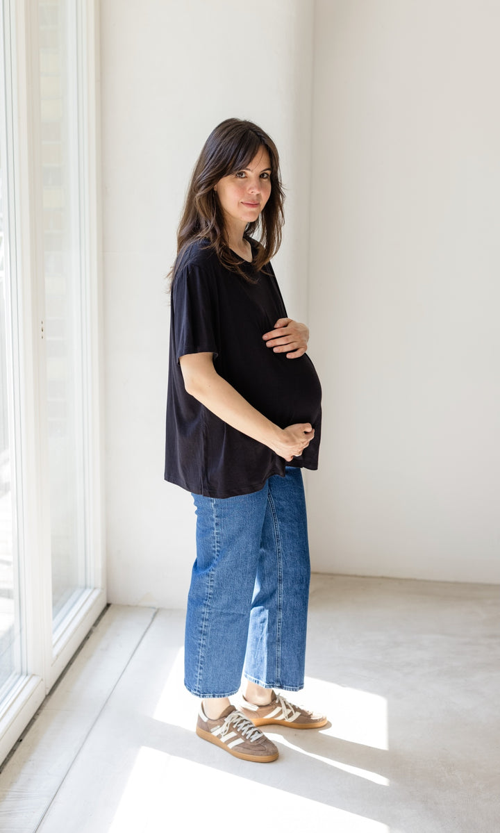 Dunkelblaue Schwangerschaftsjeans im French Fit Look mit Bauchbund kombiniert mit schwarzem T-Shirt und braunen Sneakers. Blick seitlich.