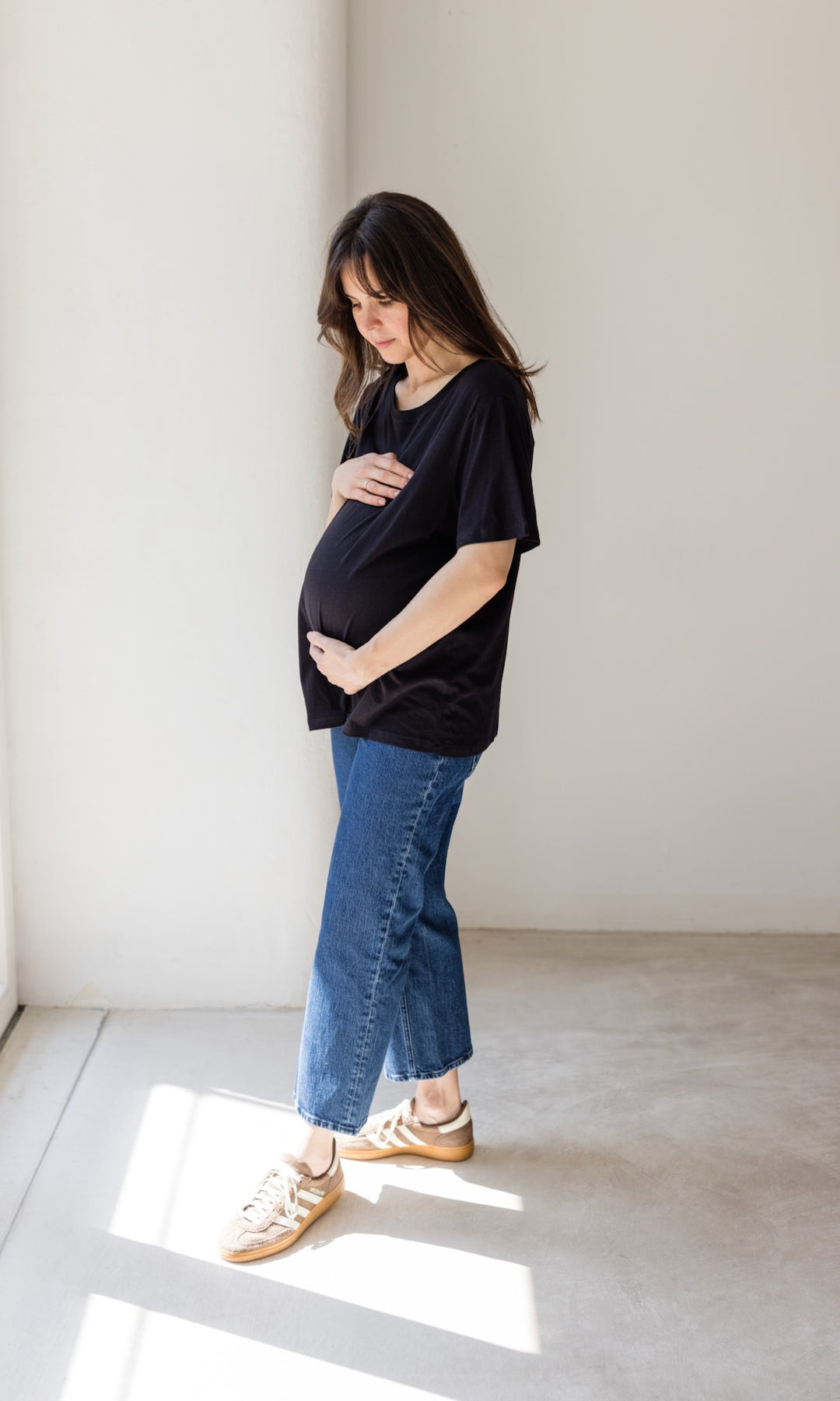 Schwangerschaftsjeans im French Fit Look in dunkelblau mit Bauchbund kombiniert mit schwarzem T-Shirt und braunen Sneakers. Blick auf Babybauch.