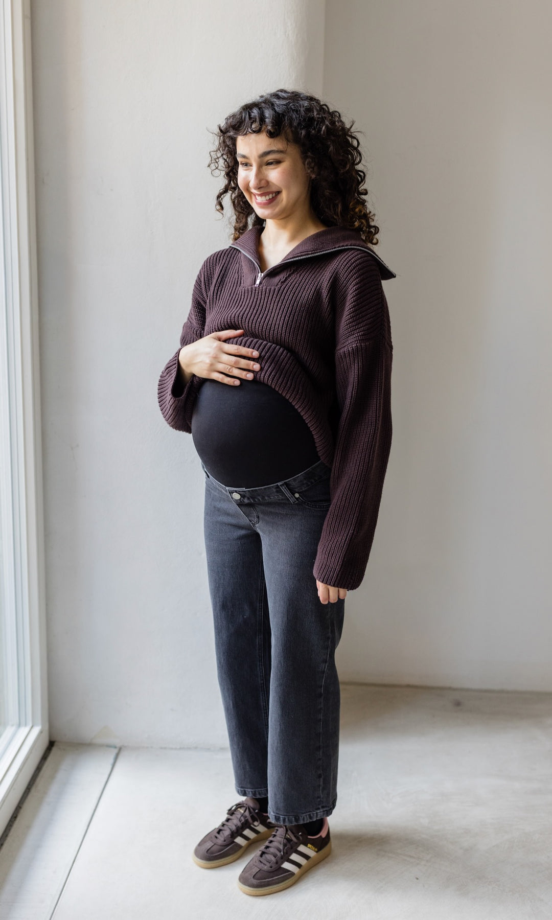 Schwarz graue Schwangerschaftsjeans im French Fit Look mit Bauchbund kombiniert mit braunem Strickpullover und braunen Sneakers. Blick gerade lachend.