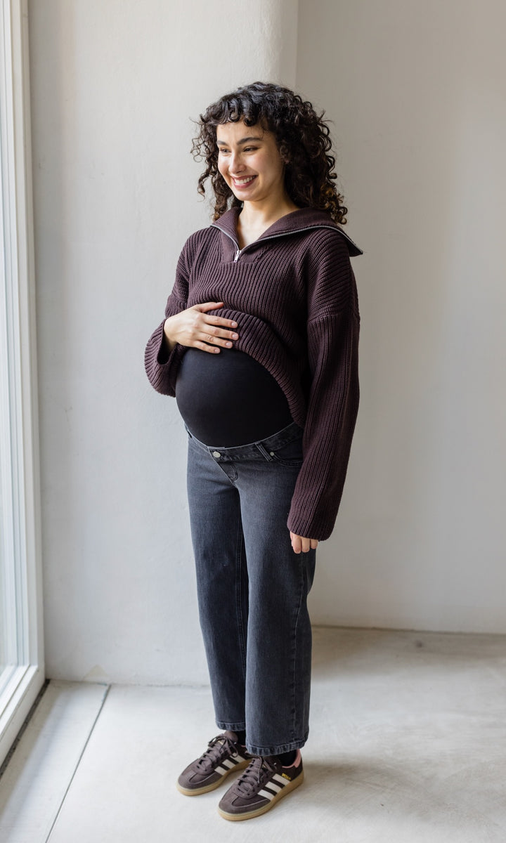 Schwarz graue Schwangerschaftsjeans im French Fit Look mit Bauchbund kombiniert mit braunem Strickpullover und braunen Sneakers. Blick gerade lachend.