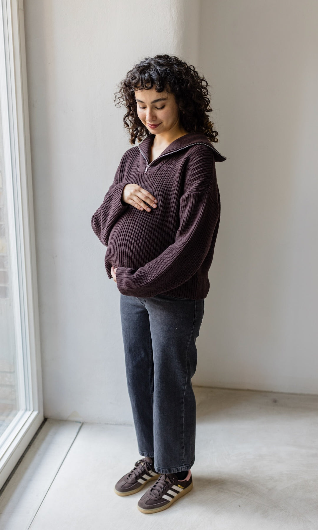 Schwangerschaftsjeans im French Fit Look in schwarz kombiniert mit braunem Strickpullover und braunen Sneakers. Inklusive elastischem Bauchbund. Blick auf Babybauch lächelnd.