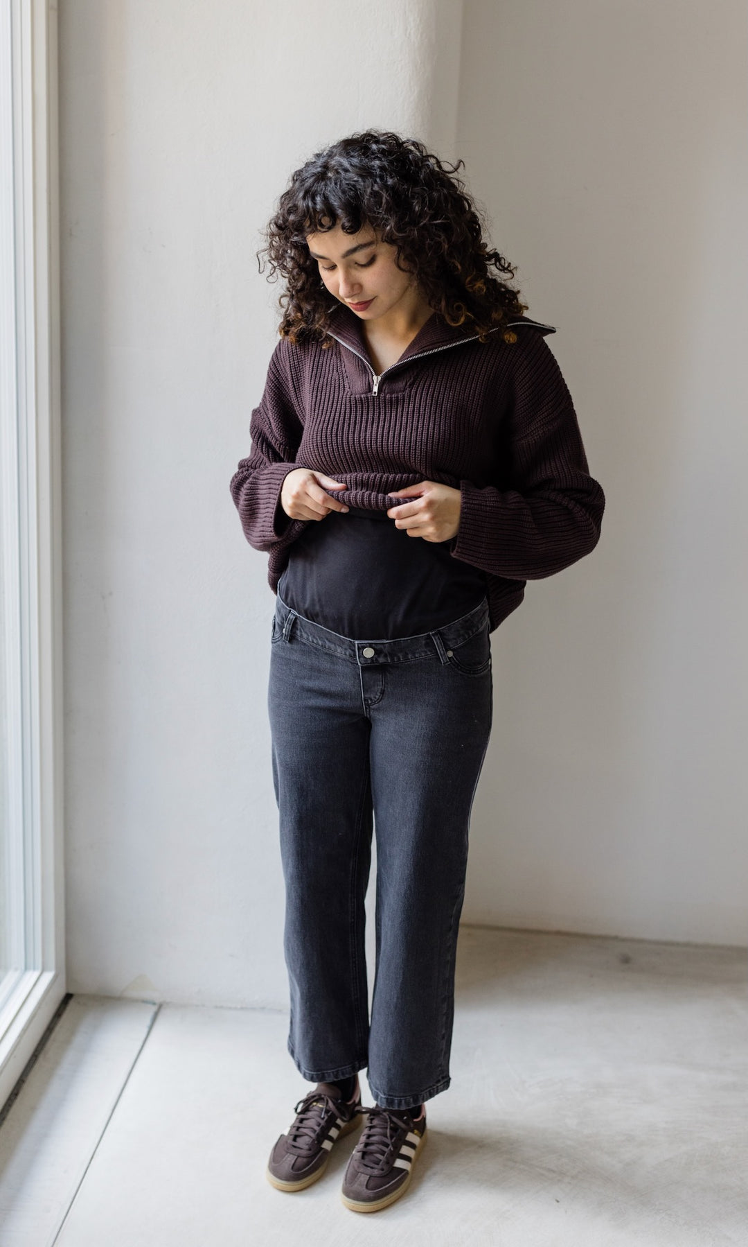 Schwarze Schwangerschaftsjeans im French Fit Look kombiniert mit braunem Strickpullover und braunen Sneakers. Blick auf elastischen Bauchbund.