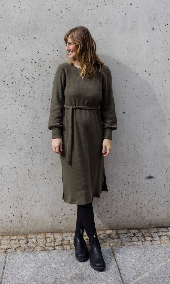 Still Strickkleid Jules in grün olive mit Stiefeln frontal vor Wand Blick seitlich