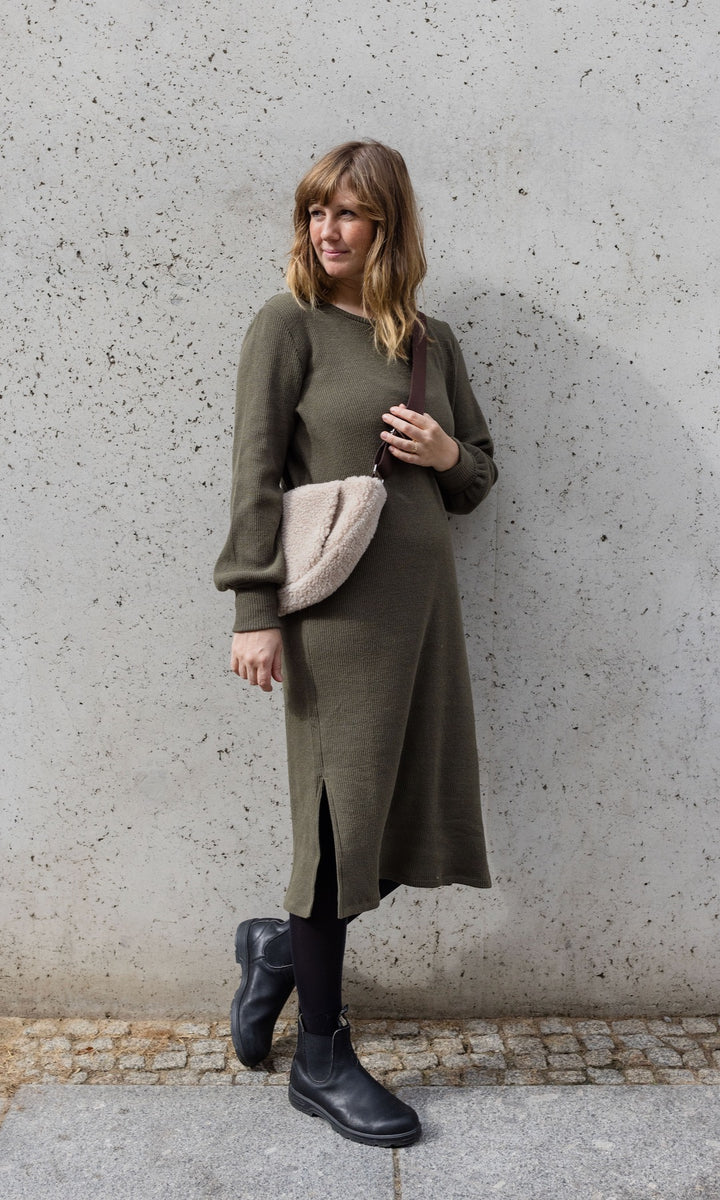 Still Strickkleid Jules in grün olive mit Stiefeln und Tasche seitlich vor Wand