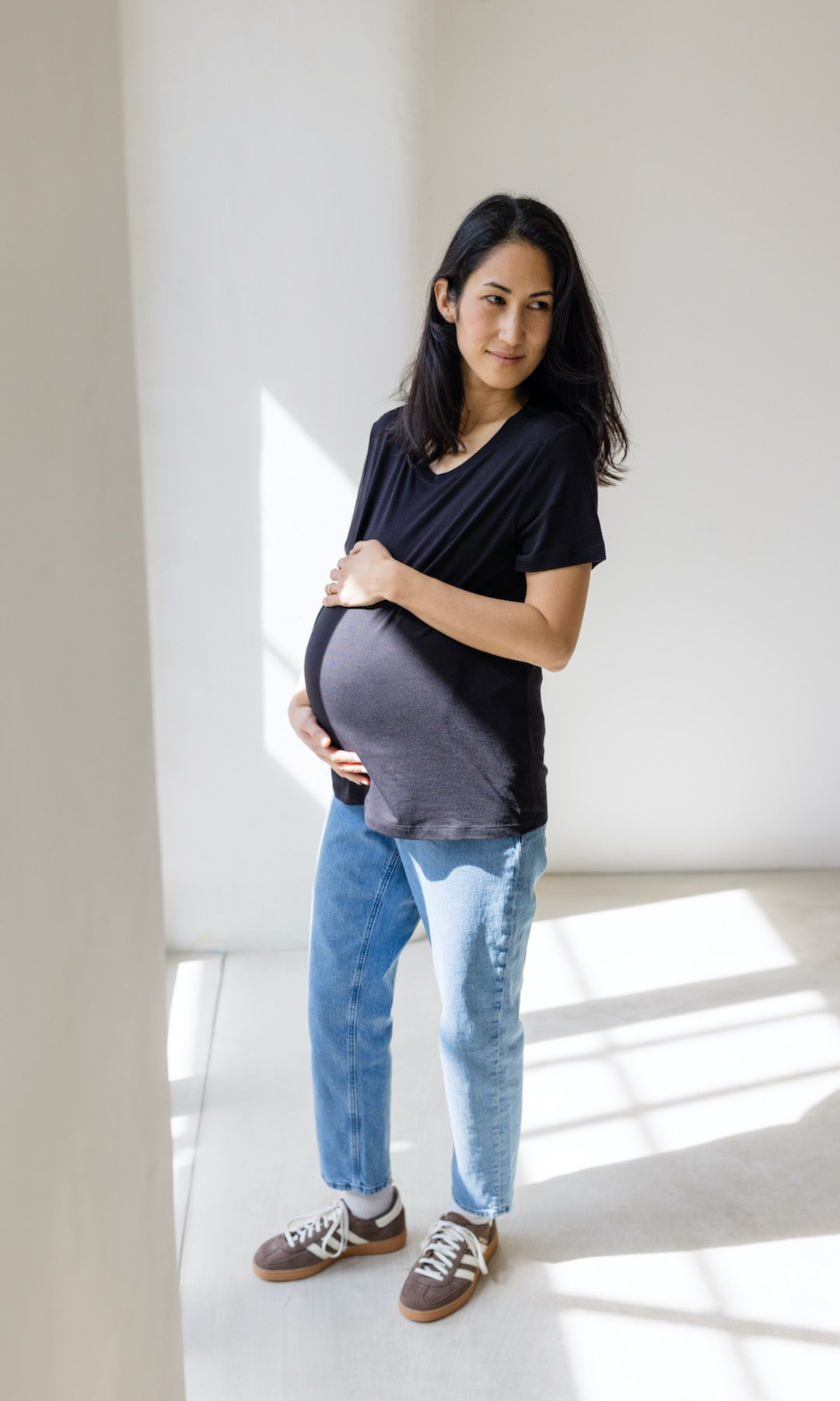Umstandsjeans im Mom Fit Look in hellblau mit Bauchbund kombiniert mit schwarzem T-Shirt und hellbraunen Sneakers. Blick seitlich lächelnd.