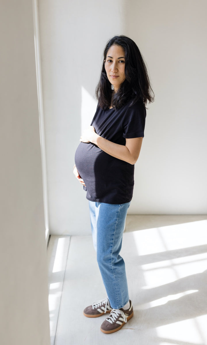 Hellblaue Umstandsjeans im Mom Fit Look mit Bauchbund kombiniert mit schwarzem T-Shirt und hellbraunen Sneakers. Blick seitlich lächelnd.