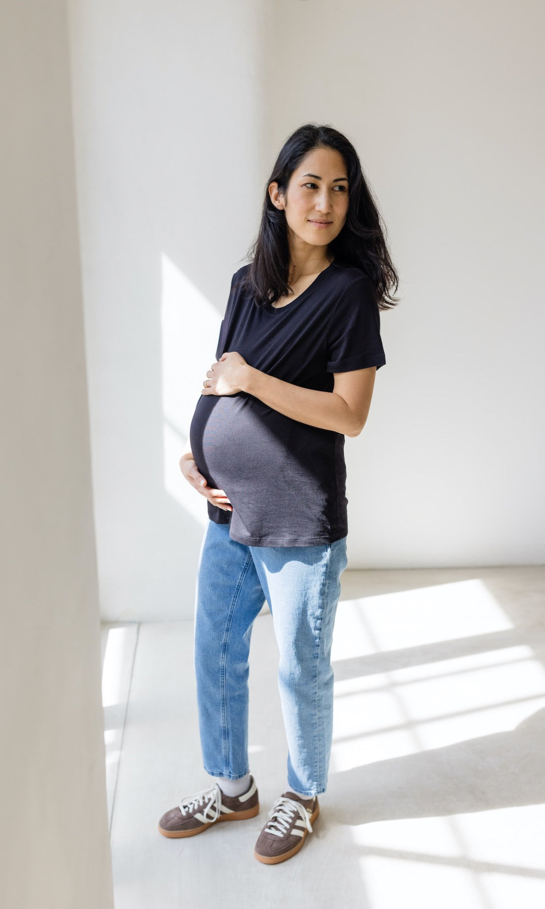 Blaue Umstandsjeans im Mom Fit Look mit Bauchbund kombiniert mit schwarzem T-Shirt und hellbraunen Sneakers. Blick seitlich lächelnd.