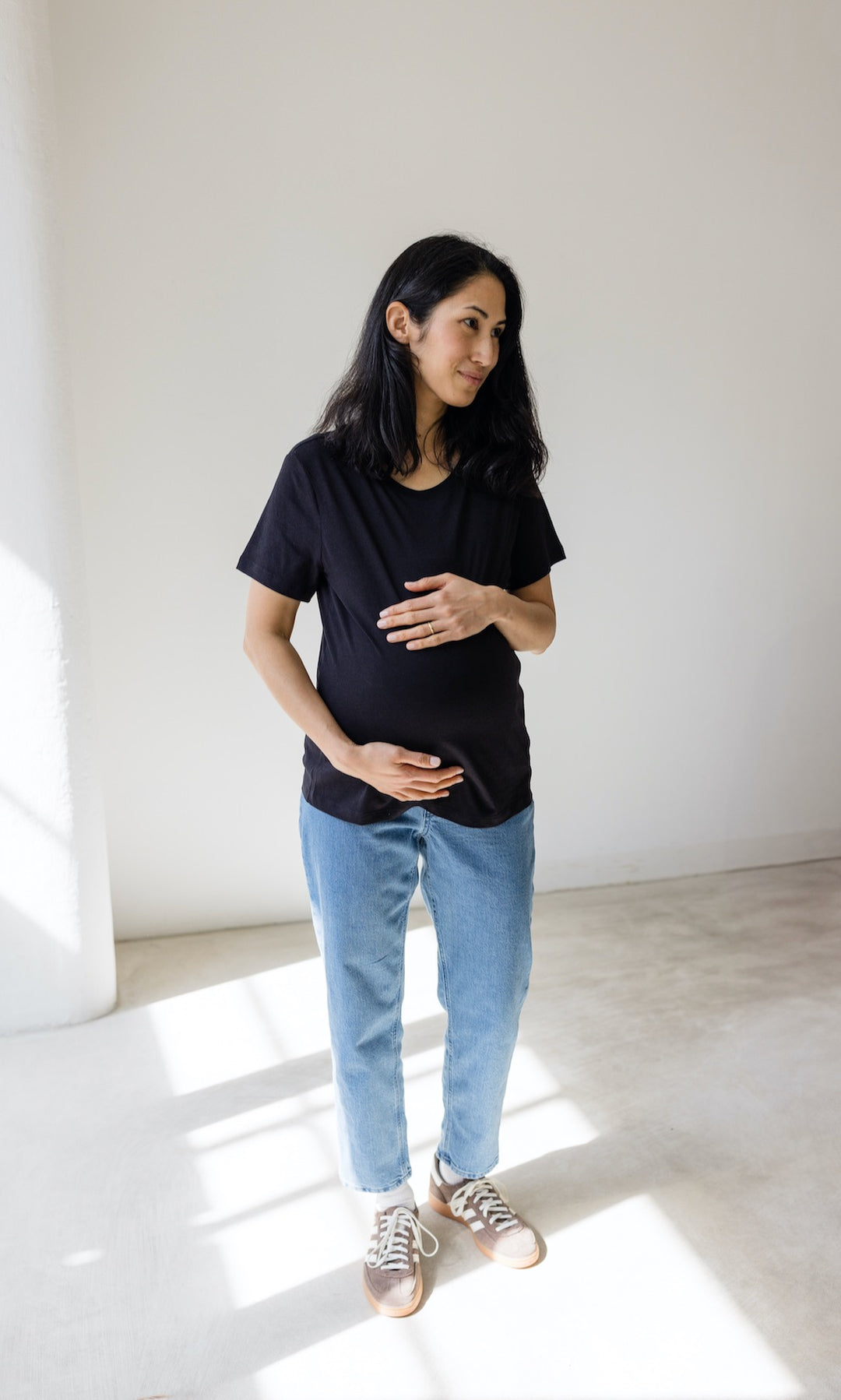 Umstandsjeans im Mom Fit Look in hellblau mit Bauchbund kombiniert mit schwarzem T-Shirt und hellbraunen Sneakers. Blick seitlich.