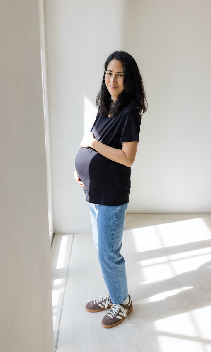 Hellblaue Umstandsjeans im Mom Fit Look mit Bauchbund kombiniert mit schwarzem T-Shirt und hellbraunen Sneakers. Seitliche Aufnahme.
