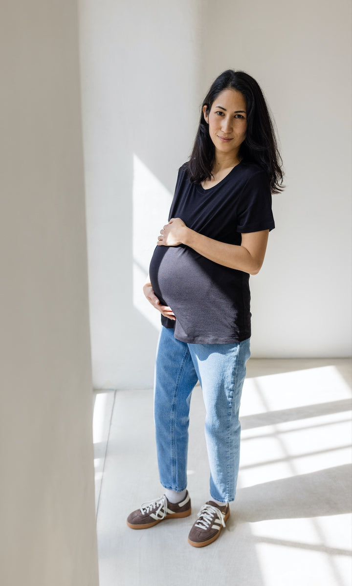 Umstandsjeans im Mom Fit Look in hellblau mit Bauchbund kombiniert mit schwarzem T-Shirt und hellbraunen Sneakers. Blick gerade.