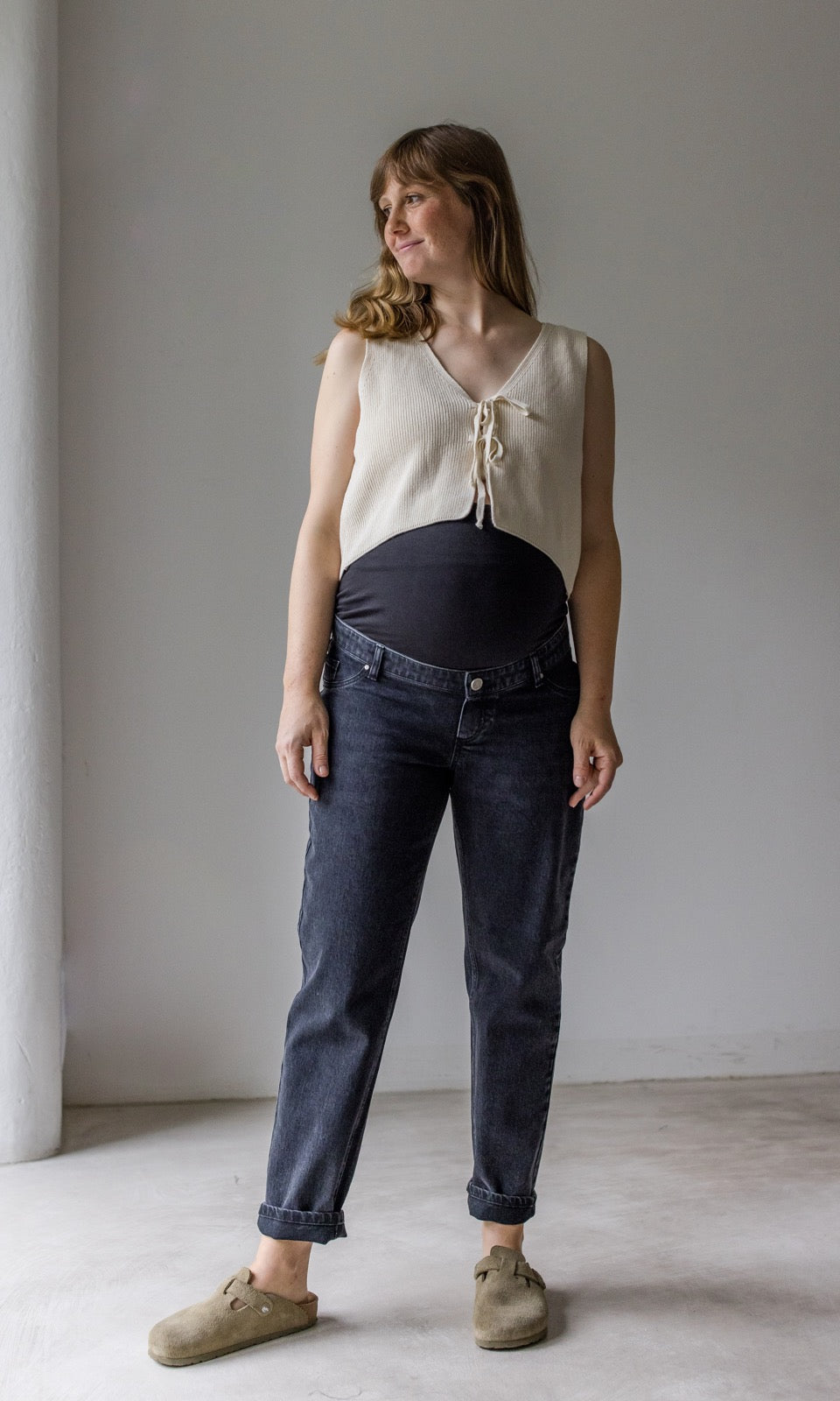 Umstandsjeans im Mom Fit Look in schwarz mit Bauchbund kombiniert mit heller Strickweste und hellbraunen Sandalen. Blick seitlich lächelnd 