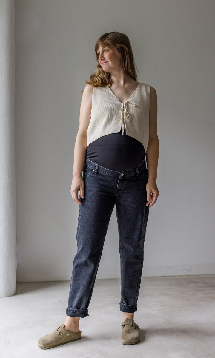 Umstandsjeans im Mom Fit Look in schwarz mit Bauchbund kombiniert mit heller Strickweste und hellbraunen Sandalen. Blick seitlich lächelnd 