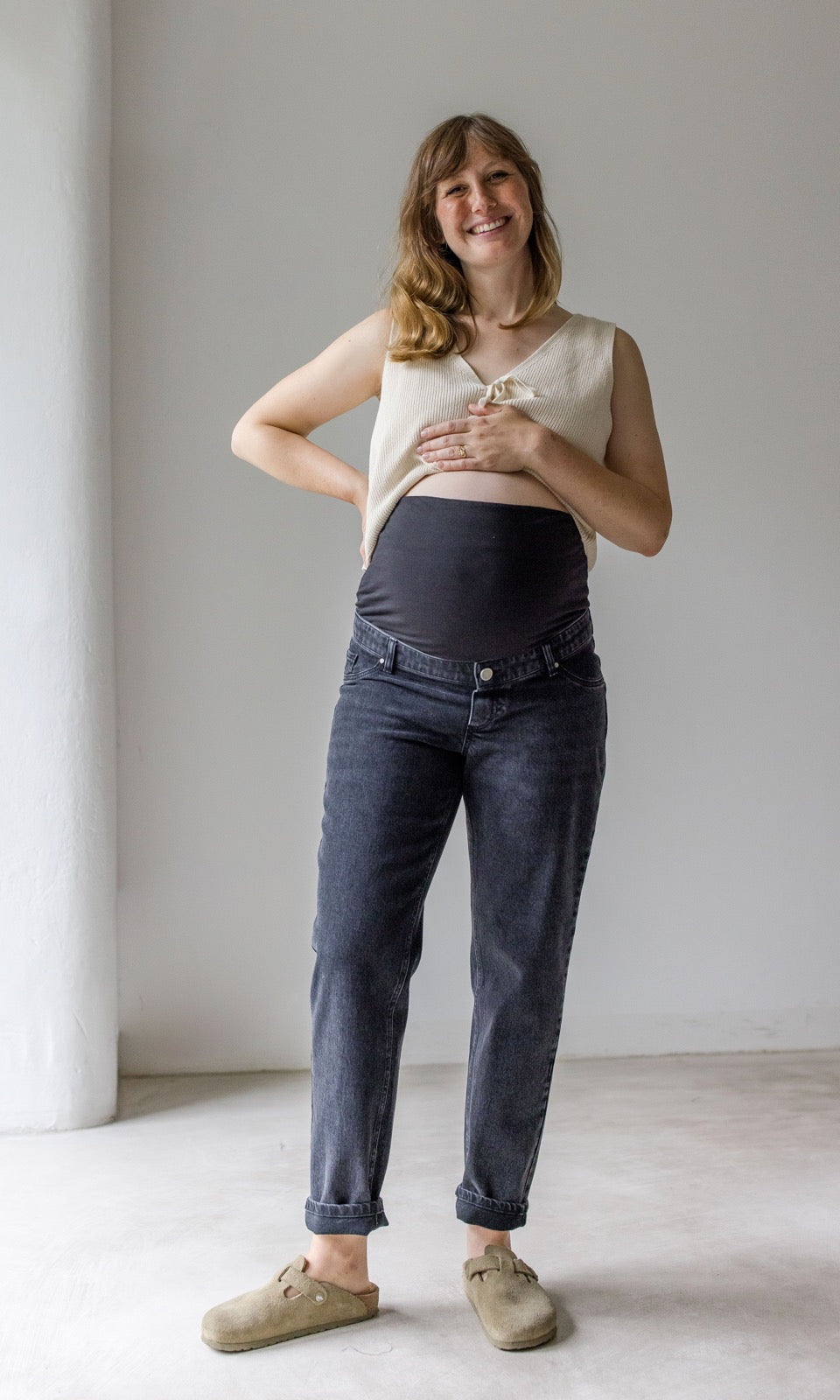 Schwarze Umstandsjeans im Mom Fit Look mit Bauchbund kombiniert mit heller Strickweste und hellbraunen Sandalen. Präsentation Bauchbund. Blick gerade lachend.