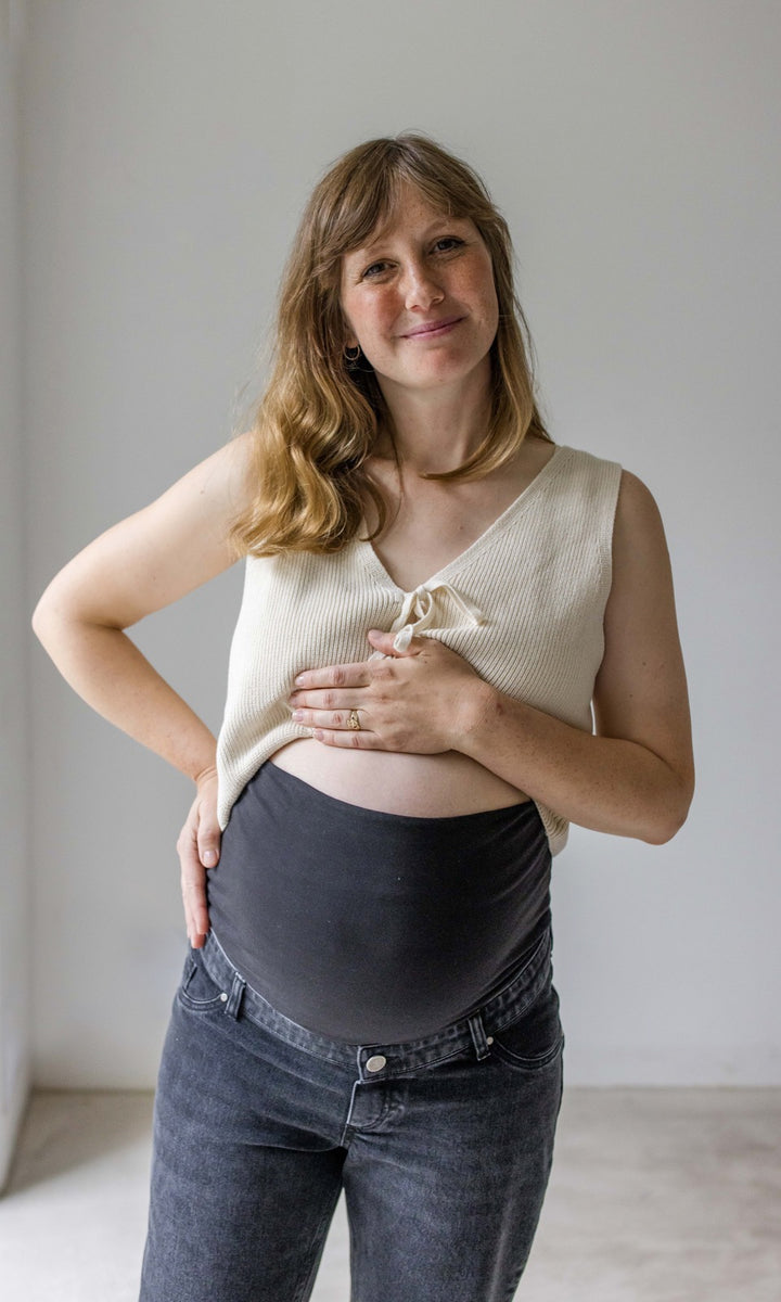 Schwarze Umstandsjeans im Mom Fit Look mit Bauchbund kombiniert mit heller Strickweste. Detailaufnahme Bauchbund. Blick gerade.