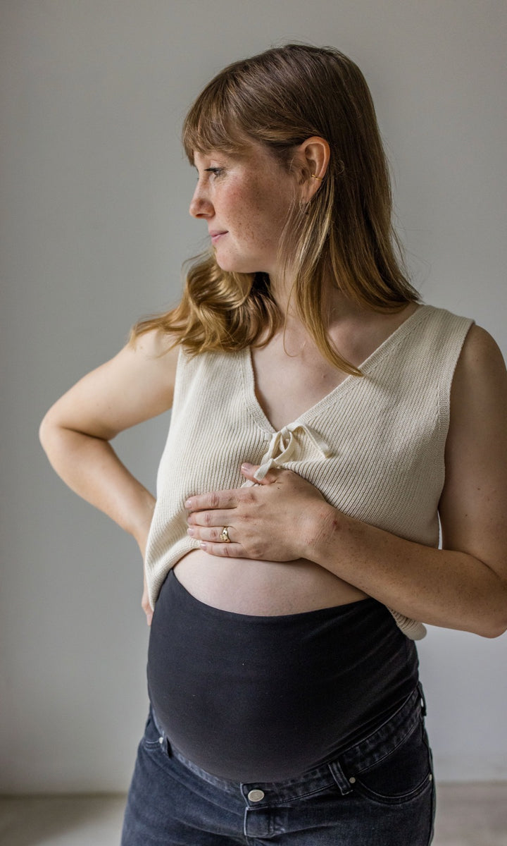 Umstandsjeans schwarz im Mom Fit Look mit Bauchbund kombiniert mit heller Strickweste. Detailaufnahme Bauchbund. Blick seitlich.