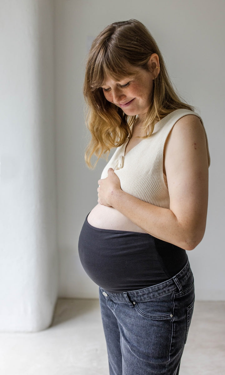 Schwarze Umstandsjeans im Mom Fit Look mit Bauchbund kombiniert mit heller Strickweste. Detailaufnahme Bauchbund. Aufnahme seitlich.
