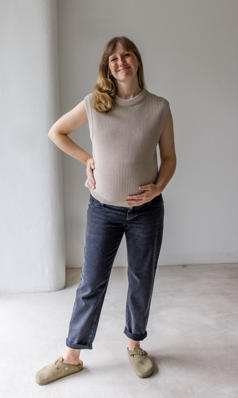 Schwarze Umstandsjeans im Mom Fit Look mit Bauchbund kombiniert mit beigem Strickpullunder und hellbraunen Sandalen. Blick gerade lächelnd.