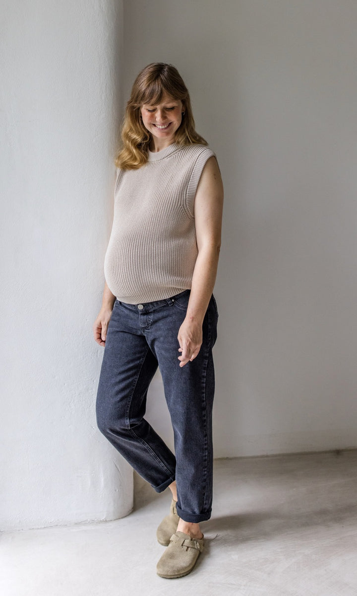 Umstandsjeans schwarz im Mom Fit Look mit Bauchbund kombiniert mit beigem Strickpullunder und hellbraunen Sandalen. Blick nach unten lachend.