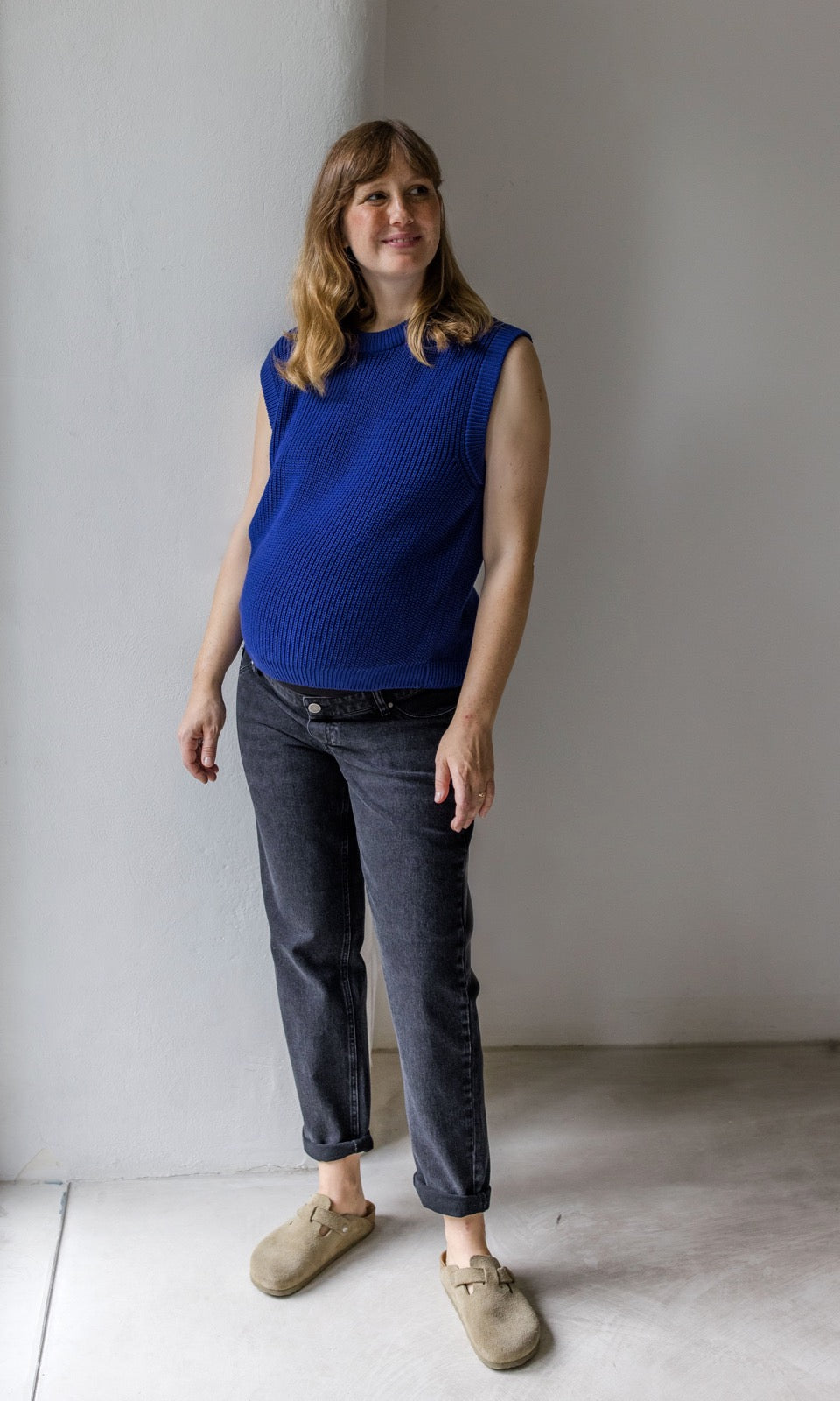 Schwarze Umstandsjeans im Mom Fit Look mit Bauchbund kombiniert mit blauem Strickpullunder und hellbraunen Sandalen. Blick seitlich lächelnd.