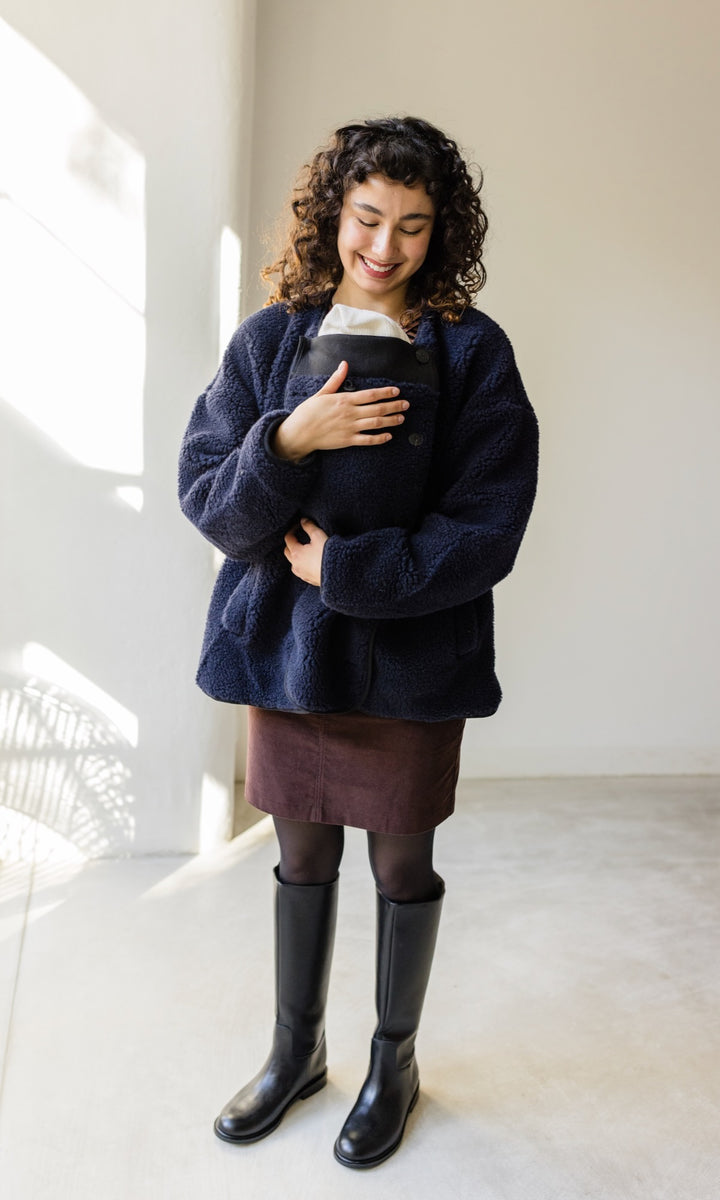 Babytrage Einsatz für Schafwoll Jacke in blau mit braunem Rock schwarzen Stiefeln vor heller Wand Blick zum Kind