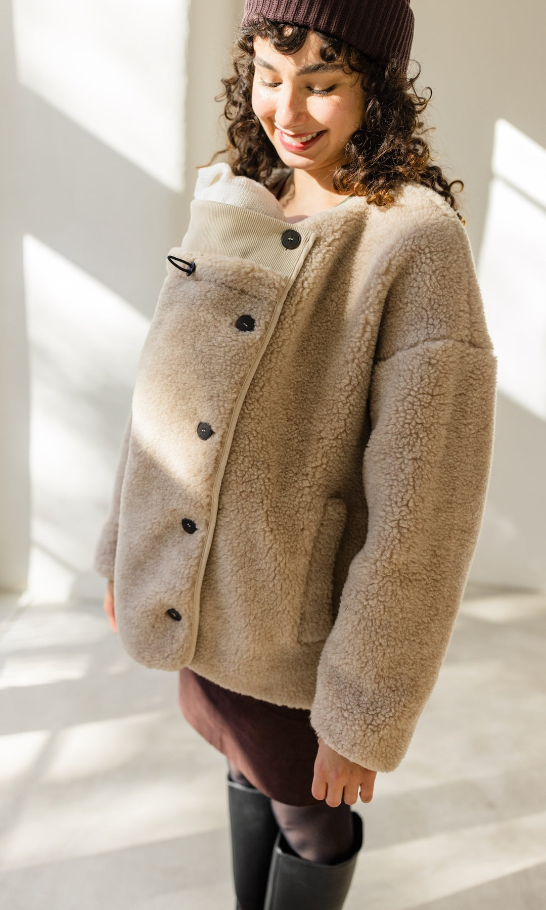 Babytrage Einsatz für Schafwoll Jacke in creme natur mit braunem Rock brauener Strickmütze vor heller Wand Blick zum Kind