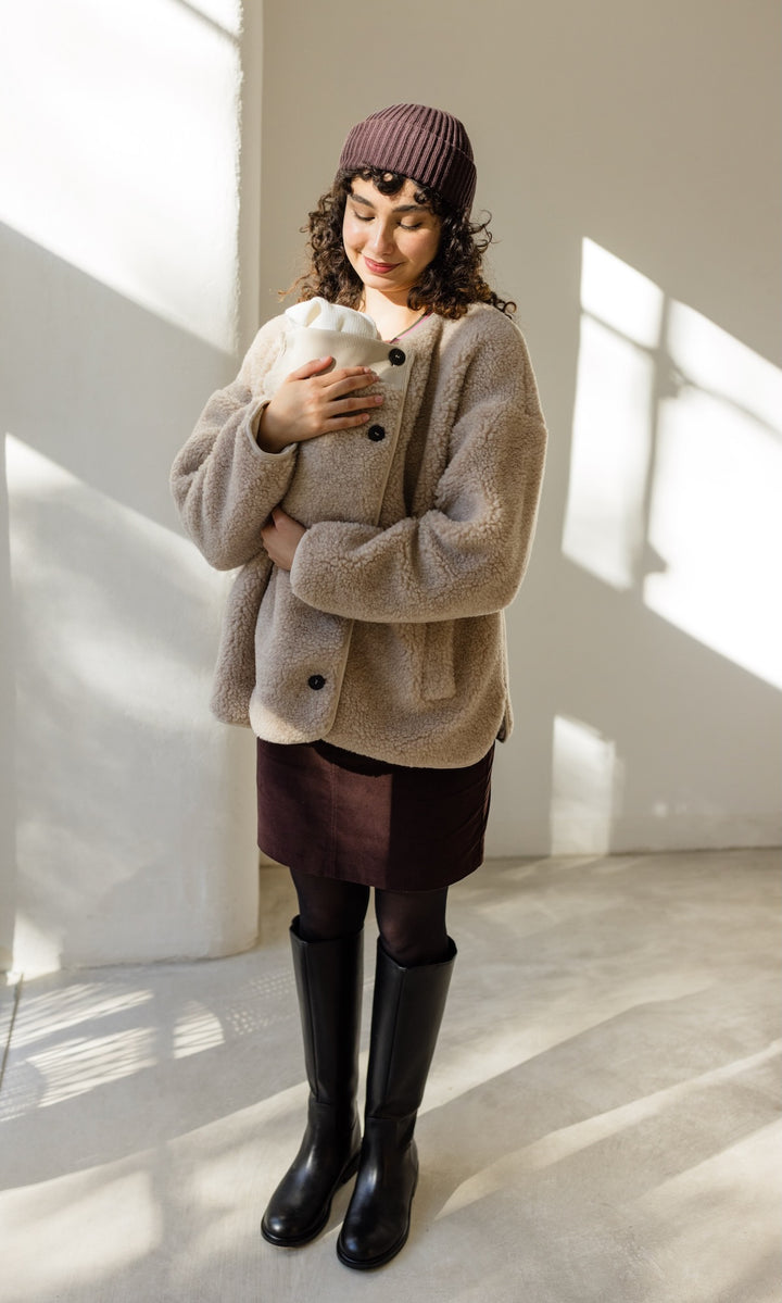 Babytrage Einsatz für Schafwoll Jacke in creme natur mit braunem Rock brauener Strickmütze und schwarzen Stiefeln vor heller Wand Blick zum Kind