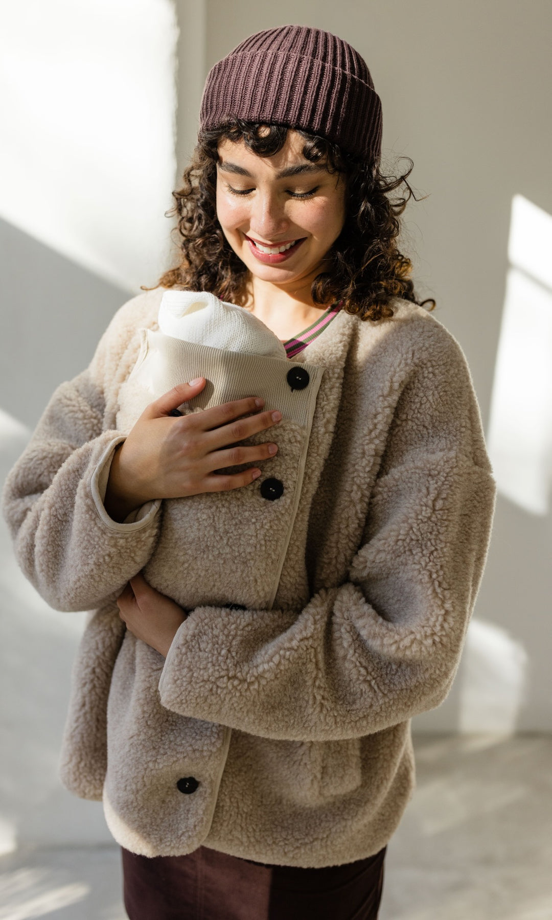 Babytrage Einsatz für Schafwoll Jacke in creme natur mit braunem Rock brauener Strickmütze vor heller Wand Blick zum Kind lächelnd