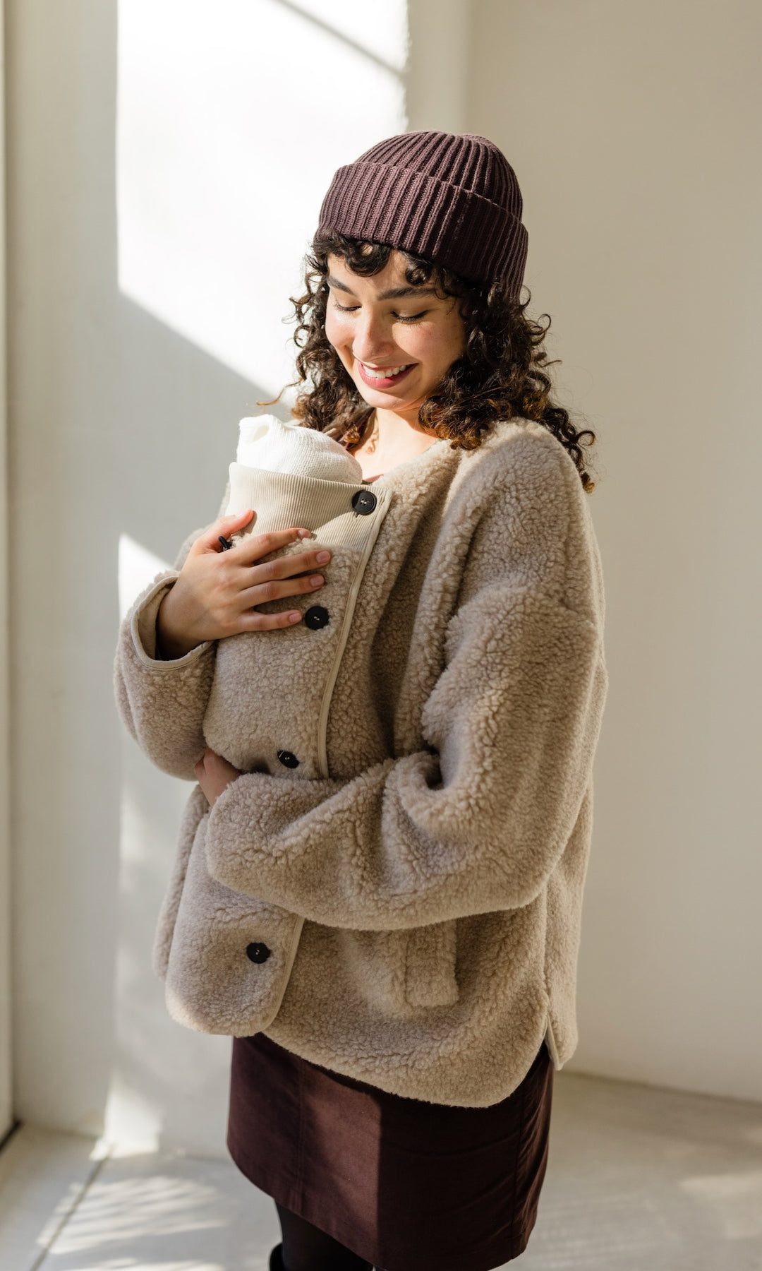 Babytrage Einsatz für Schafwoll Jacke in creme natur mit braunem Rock brauener Strickmütze vor heller Wand Blick zum Kind lächelnd