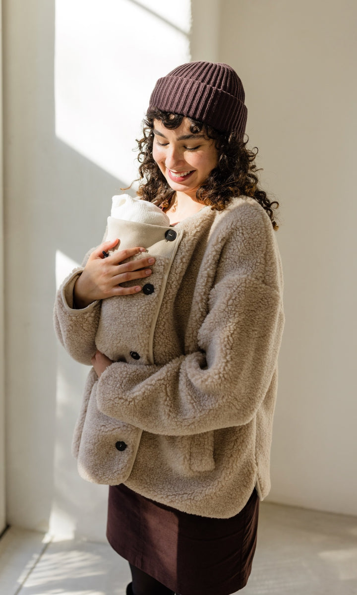 Babytrage Einsatz für Schafwoll Jacke in creme natur mit braunem Rock brauener Strickmütze vor heller Wand Blick zum Kind lächelnd