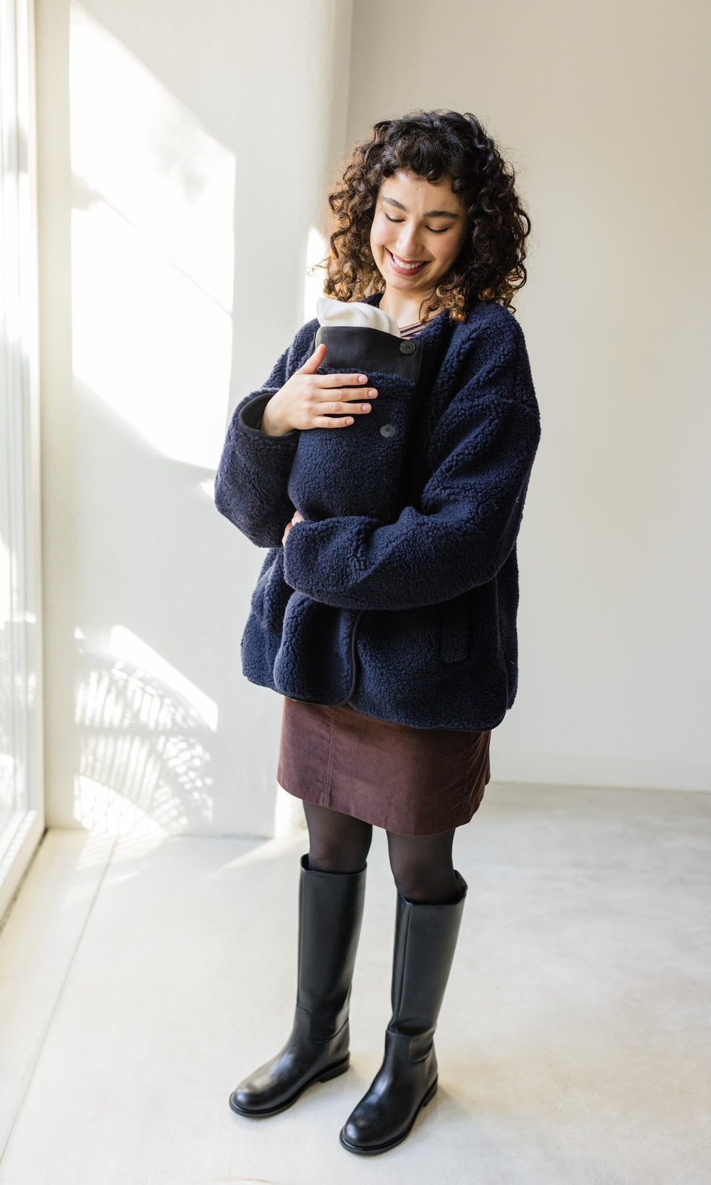 Babytrage Einsatz für Schafwoll Jacke in dunkelblau mit braunem Rock schwarzen Stiefeln vor heller Wand Blick zum Kind