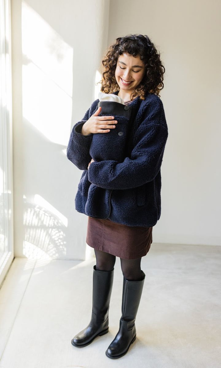 Babytrage Einsatz für Schafwoll Jacke in dunkelblau mit braunem Rock schwarzen Stiefeln vor heller Wand Blick zum Kind