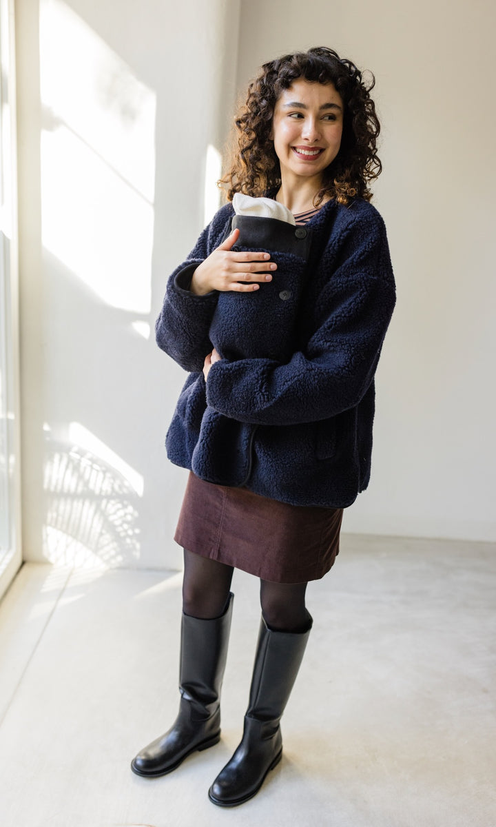 Babytrage Einsatz für Schafwoll Jacke in dunkelblau mit braunem Rock schwarzen Stiefeln vor heller Wand Blick seitlich lächelnd