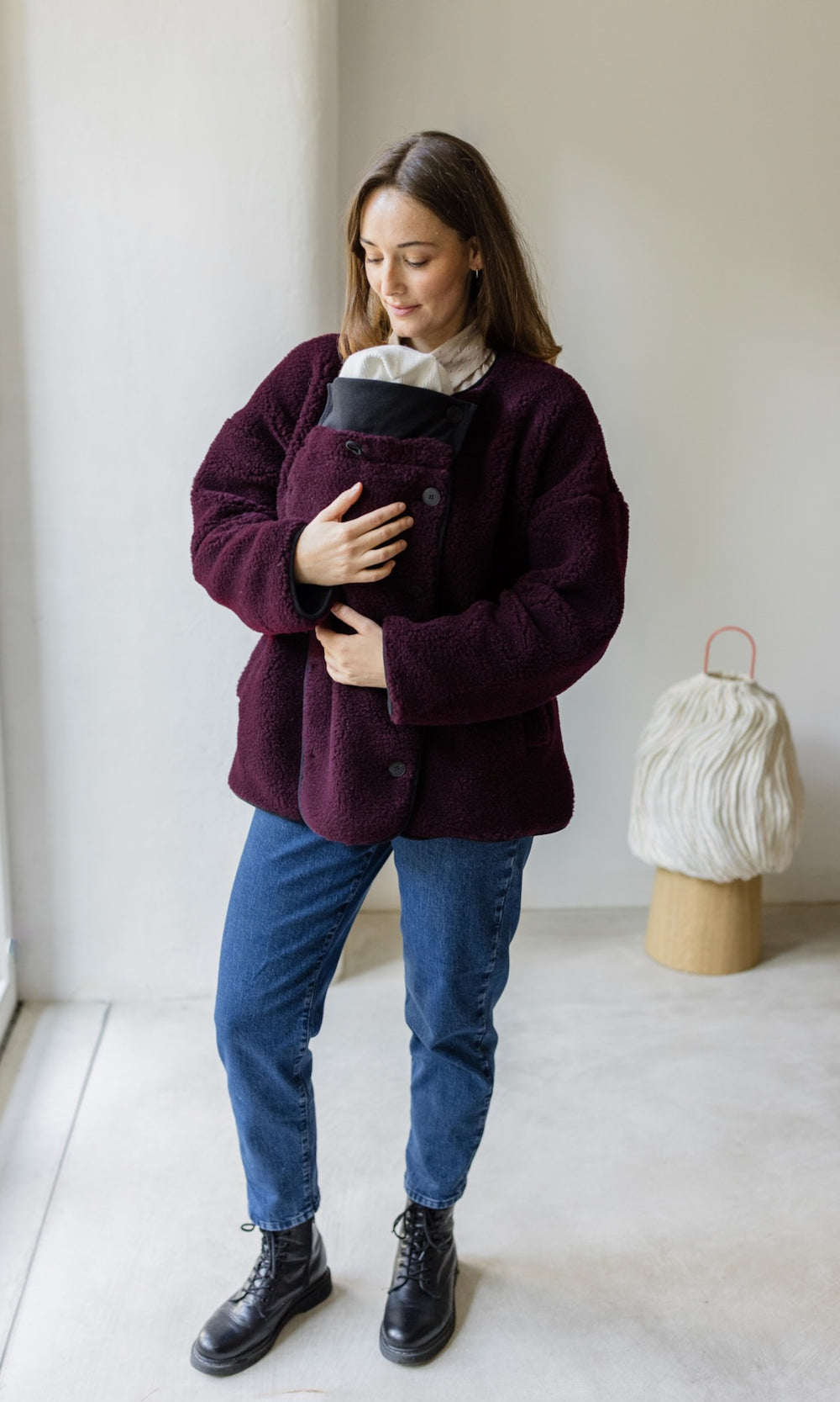 Babytrage Einsatz für Schafwoll Jacke in weinrot mit blauer Jeans und schwarzen Stiefeln vor heller Wand Blick nach unten