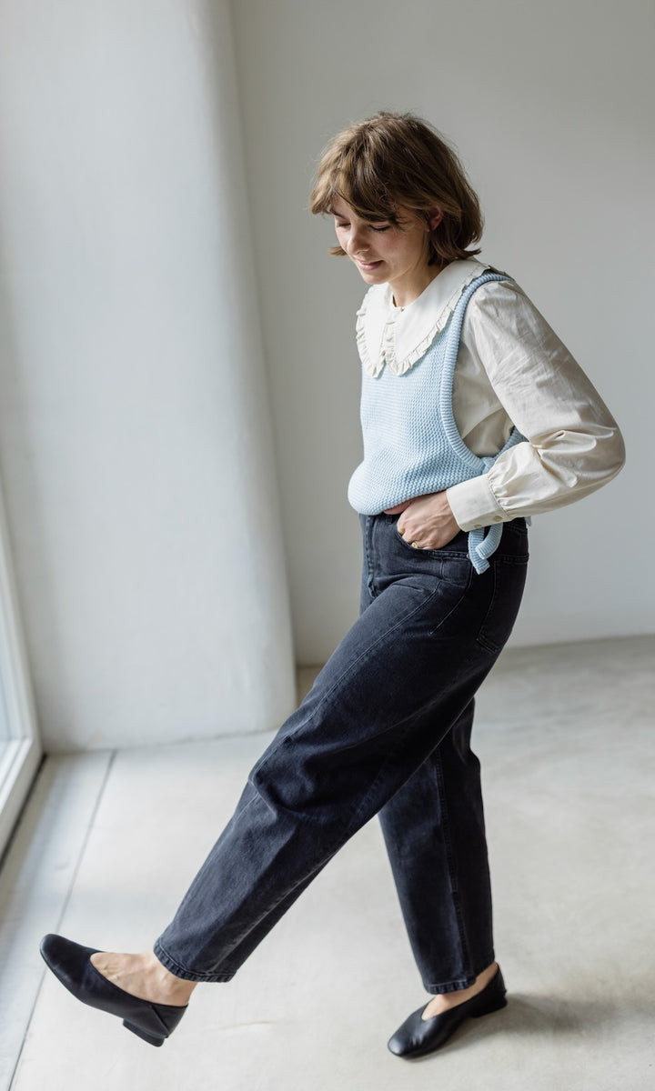 Das Modell trägt eine Barrel Jeans in schwarz kombiniert mit einem weißen, langarmigen Bluse mit großem Kragen, einem hellblauen Strickpullunder und eleganten schwarzen Loafers.