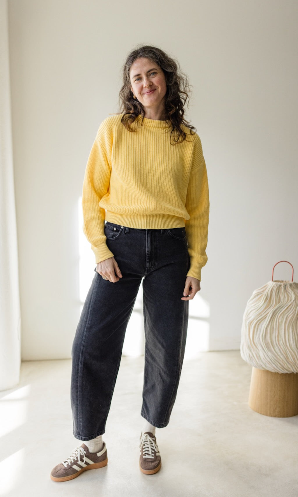 Das Modell trägt eine Barrel Jeans in schwarz kombiniert mit einem gelben Strickpullover und braunen Sneakers. Ein Casual Look für Städtereisen.