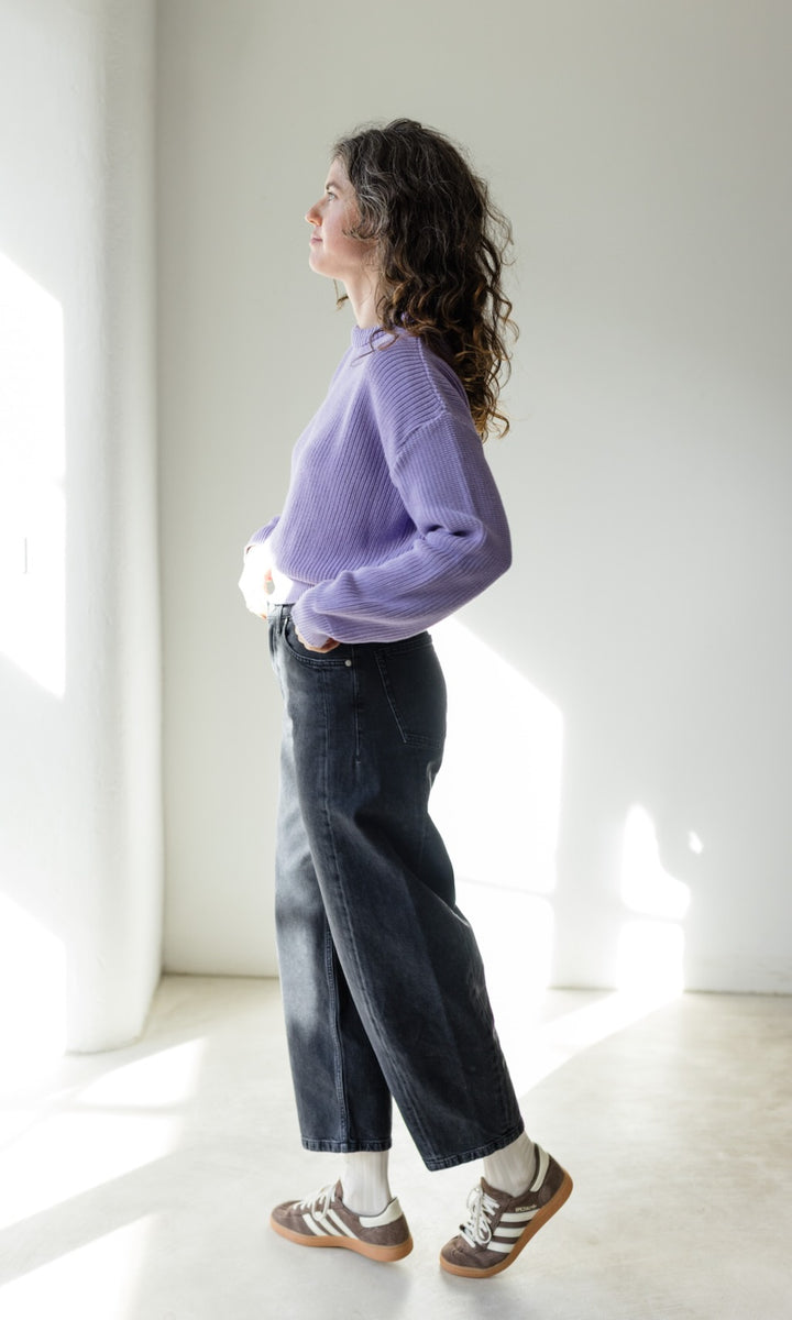 Das Modell trägt eine Barrel Jeans in schwarz kombiniert mit einem lila Strickpullover und braunen Sneakers. Ein Casual Look für Städtereisen.