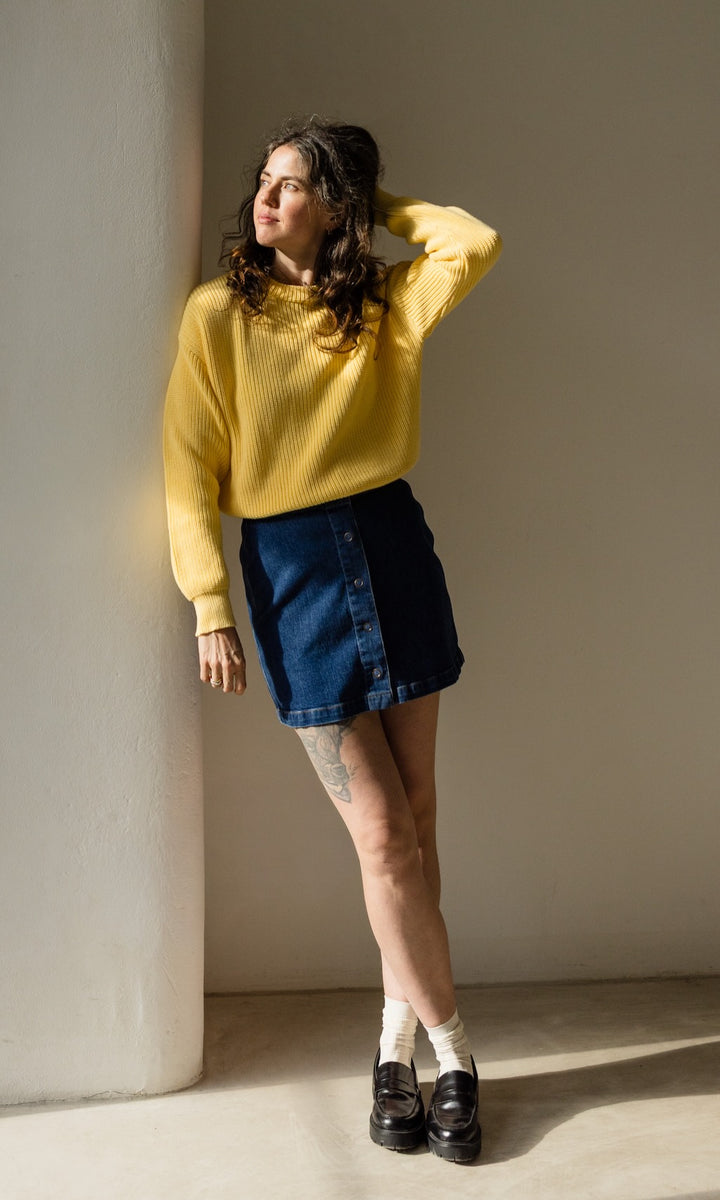 Gelber Strickpullover aus Bio-Baumwolle kombiniert mit dunkelblauem Jeansrock und schwarzen Loafers für ein lässig schickes Outfit.