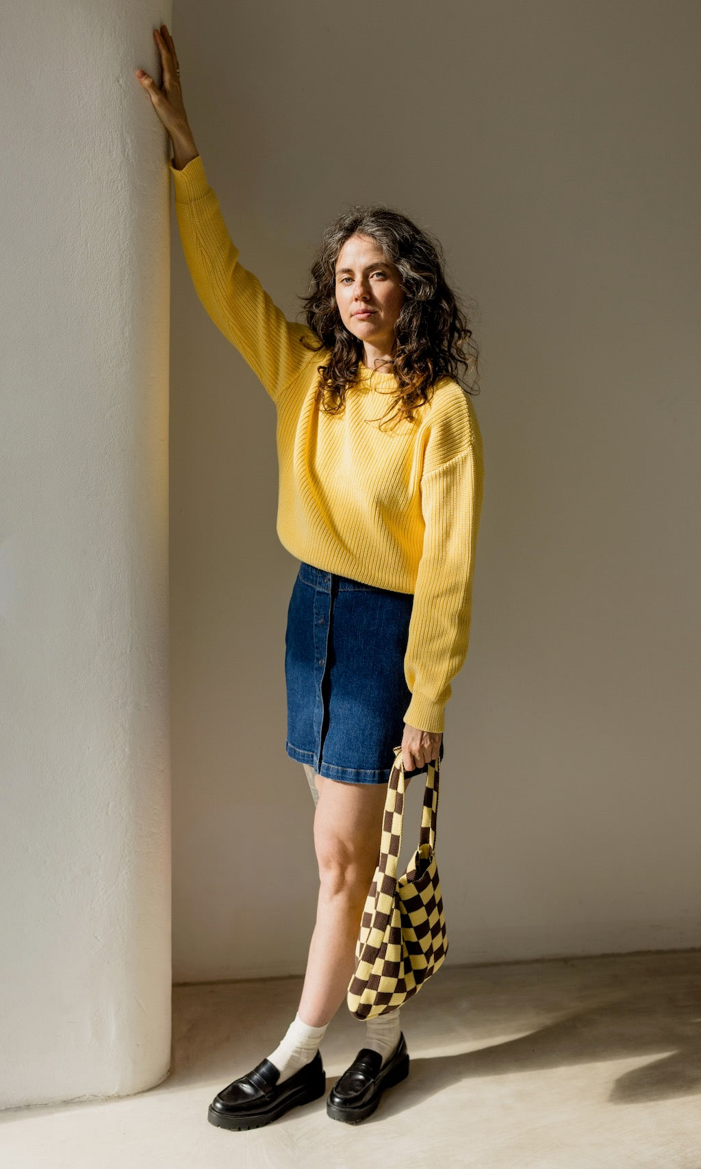 Gelber Strickpullover aus Bio-Baumwolle kombiniert mit dunkelblauem Jeansrock, gelbe/braun-karierter Stricktasche und schwarzen Loafers für ein lässig schickes Outfit.