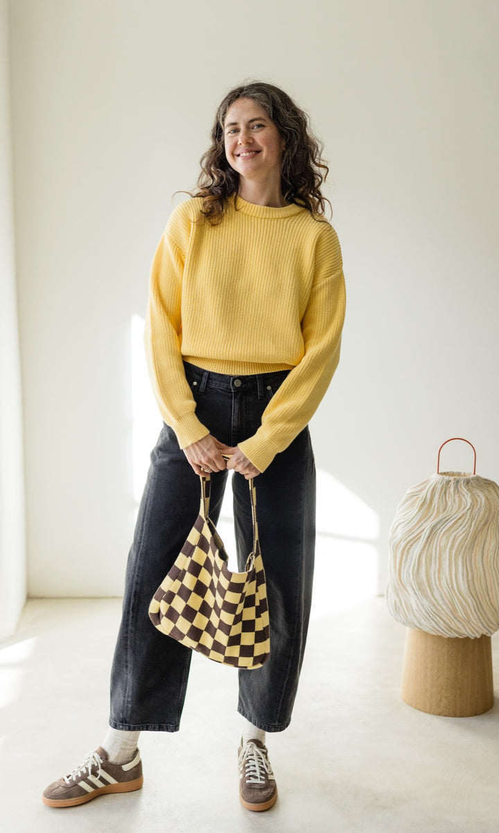 Gelber Strickpullover aus Bio-Baumwolle kombiniert mit schwarzer Barrel-Jeans, gelb/brauner Stricktasche und braunen Sneakers für ein lässiges Outfit.