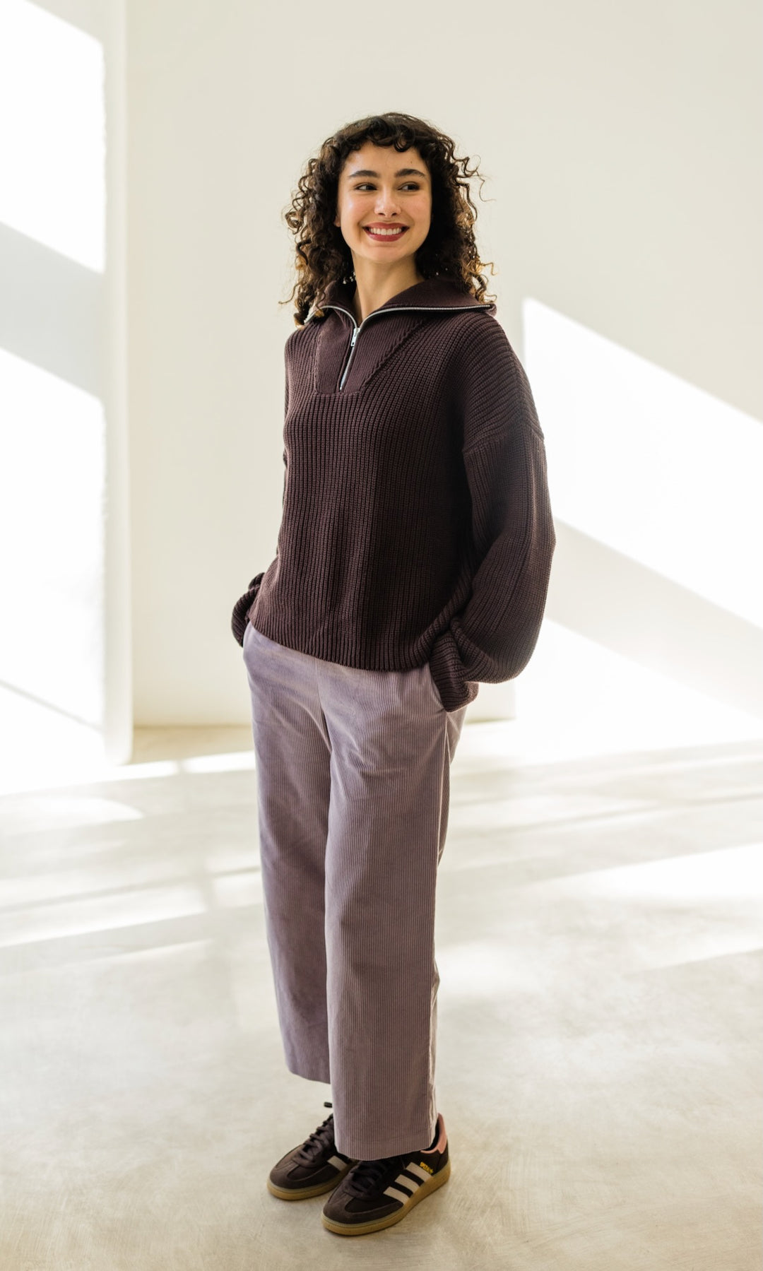 Gerippter Pulli mit Zipper Reissverschluss in braun mit hellbrauner Hose und braunen Sneakers vor heller Wand lachend