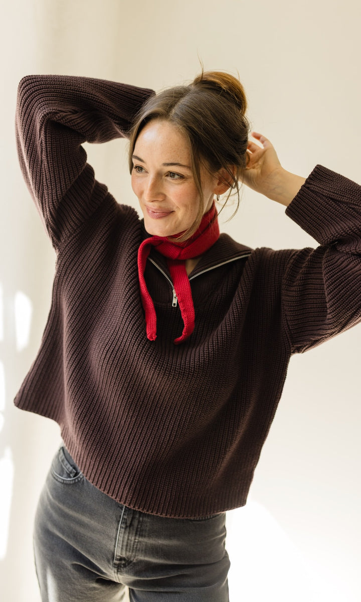 Gerippter Pulli mit Zipper Reissverschluss in braun mit dunkler Jeans und rotem kleinem Strickschal vor heller Wand Blick seitlich lächelnd